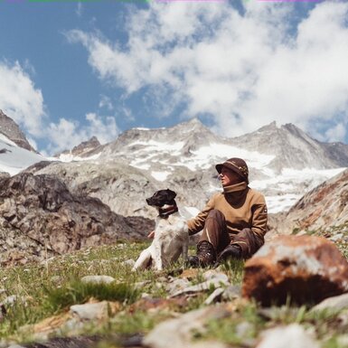 Bergwanderer sitzt inmitten einer Berglandschaft mit schneebedeckten Gipfeln und begleitet von einem Hund, umgeben von Felsen und Grünflächen unter einem wolkenverhangenen Himmel. | © Urlaub am Bauernhof / Daniel Gollner