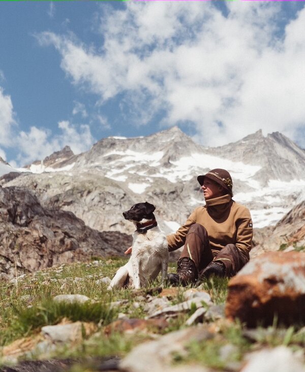 Bergwanderer sitzt inmitten einer Berglandschaft mit schneebedeckten Gipfeln und begleitet von einem Hund, umgeben von Felsen und Grünflächen unter einem wolkenverhangenen Himmel. | © Urlaub am Bauernhof / Daniel Gollner