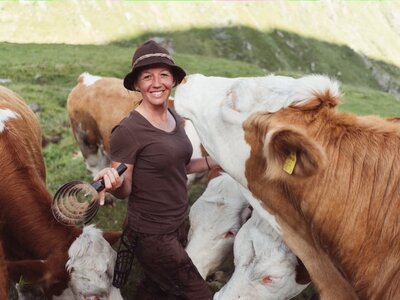 Eine Frau in Arbeitskluft und Hut lächelt umgeben von mehreren Kühen auf einer grünen Wiese. Sie melkt eine Kuh manuell mit einem Melkeimer. | © Urlaub am Bauernhof / Daniel Gollner