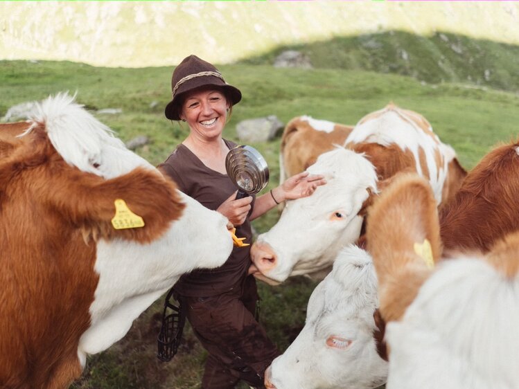 Lächelnde Frau in Arbeitskleidung umgeben von einer Gruppe bunter Kühe auf einer grünen Wiese mit Bergen im Hintergrund.