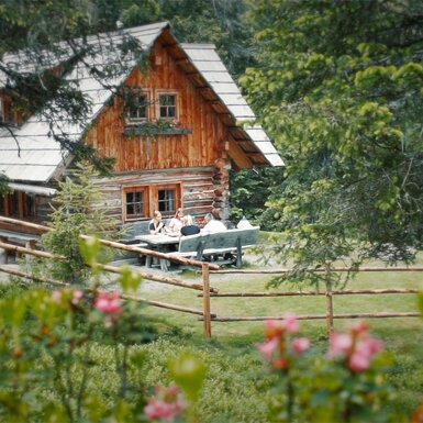 Malerisches Holzhaus umgeben von Bäumen und Blumen, auf einer Terrasse sitzen Menschen und genießen die Natur.