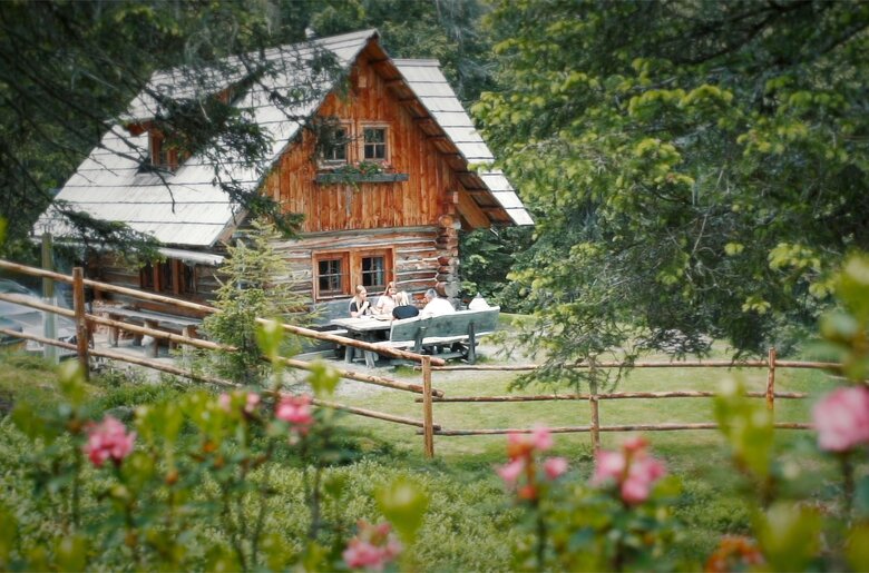 Malerisches Holzhaus umgeben von Bäumen und Blumen, auf einer Terrasse sitzen Menschen und genießen die Natur.