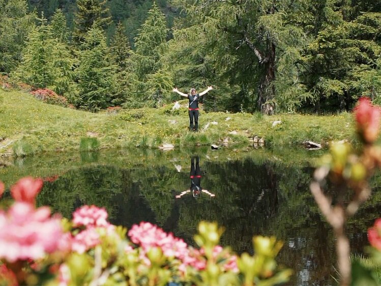 Person mit ausgebreiteten Armen steht in einem idyllischen See, umgeben von üppigem Grün und bunten Blüten. Die Spiegelung der Person im Wasser schafft einen ruhigen und harmonischen Gesamteindruck.