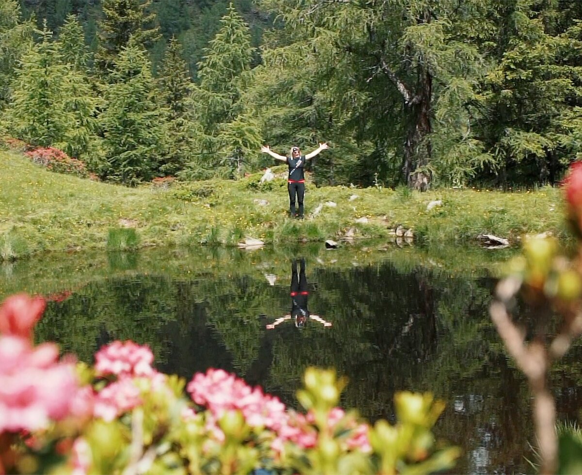 Person mit ausgebreiteten Armen steht in einem idyllischen See, umgeben von üppigem Grün und bunten Blüten. Die Spiegelung der Person im Wasser schafft einen ruhigen und harmonischen Gesamteindruck.