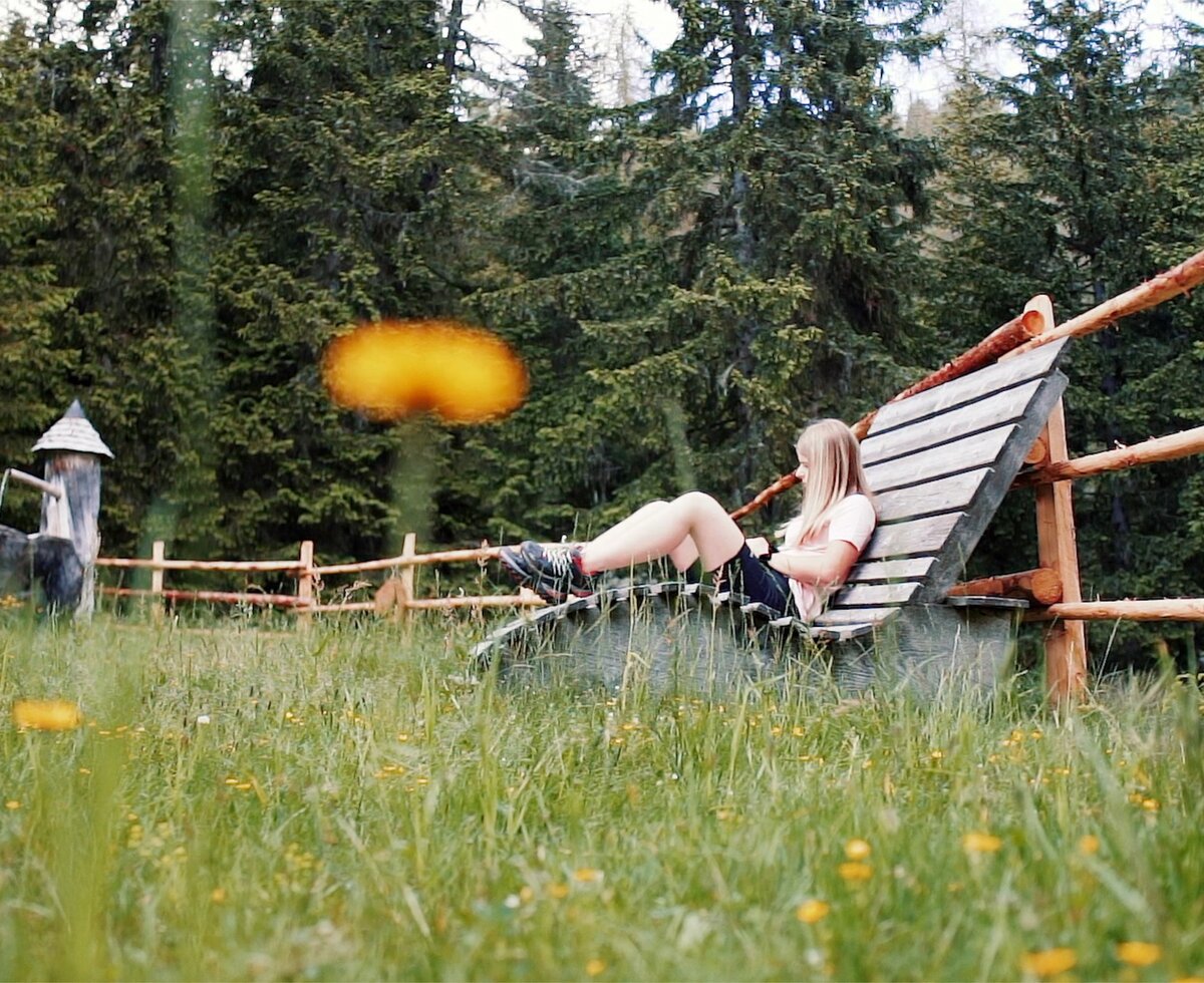 Eine Person liegt entspannt auf einer hölzernen Bank, umgeben von waldiger Landschaft und blühenden Blumenwiesen. Eine pittoreske ländliche Szene mit einem Spielturm im Hintergrund.