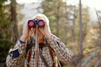 Ein Person mit Fernglas beobachtet aufmerksam die umgebende Natur im Wald. Die Person trägt ein kariertes Hemd und hält die Ferngläser fest vor ihr Gesicht. | © Urlaub am Bauernhof Kärnten / Daniel Gollner