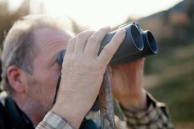 Ein älterer Mann begutachtet die Umgebung konzentriert durch ein Fernglas im Freien. | © Urlaub am Bauernhof Kärnten / Daniel Gollner