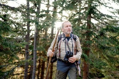 Ein älterer Mann mit Rucksack und Kamera, umgeben von Bäumen und Natur, inmitten eines Waldes mit Sonneneinstrahlung. Er scheint zu wandern und die Umgebung zu erkunden. | © Urlaub am Bauernhof Kärnten / Daniel Gollner