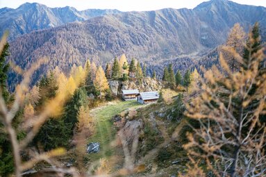 Malerische Berglandschaft mit herbstlichem Laubwald, einem Bergbach und rustikalen Holzhäusern auf einer Bergkuppe. | © Urlaub am Bauernhof Kärnten / Daniel Gollner