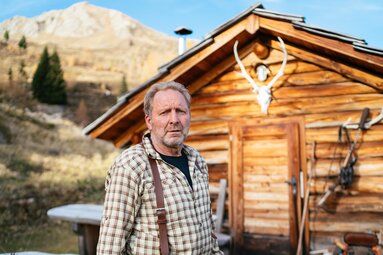 Ein älterer Mann mit Karohemd und Hosenträgern steht vor einer traditionellen Holzhütte in einer bergigen Landschaft. | © Urlaub am Bauernhof Kärnten / Daniel Gollner