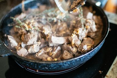 Eine dampfende Bratpfanne mit geschmortem Fleisch, umgeben von Gewürzen und Aromen. Die Zubereitung erscheint köstlich und sorgfältig. | © Urlaub am Bauernhof Kärnten / Daniel Gollner