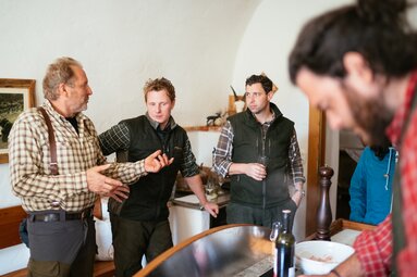 Vier Männer in karierter und grüner Kleidung stehen in einem Raum mit Bildern an den Wänden und diskutieren miteinander. | © Urlaub am Bauernhof Kärnten / Daniel Gollner