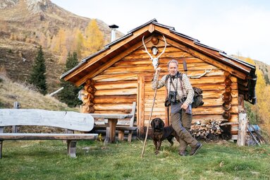 Holzblockhaus im ländlichen Bereich mit Mann und Hund davor, umgeben von Bergen und Herbstlandschaft. Der Mann trägt Bergtourenausrüstung und posiert vor dem traditionellen Alpenhäuschen. | © Urlaub am Bauernhof Kärnten / Daniel Gollner