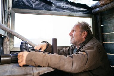Alter Mann sieht nachdenklich aus dem Fenster eines alten Wohnwagens. Seine Hände ruhen auf dem Lenkrad, umgeben von Vorhängen und Belüftung. | © Urlaub am Bauernhof Kärnten / Daniel Gollner