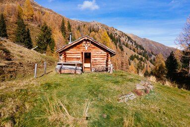 Eine rustikale Berghütte inmitten von Wiesen und Wäldern mit herbstlichen Farben, vor majestätischer Bergkulisse, lädt zum Verweilen und Genießen der Natur ein. | © Urlaub am Bauernhof Kärnten / Daniel Gollner