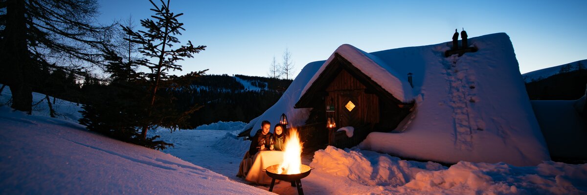 Eine verschneite Berghütte in der Abenddämmerung, vor der ein Lagerfeuer brennt und Menschen in gemütlicher Atmosphäre sitzen. | © Urlaub am Bauernhof Kärnten / Daniel Gollner