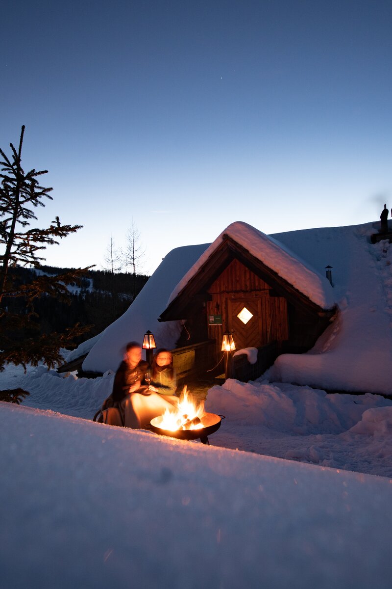 Schneebedecktes Holzhäuschen mit erleuchteten Fenstern und Lagerfeuer im Vordergrund, umgeben von verschneiter Winterlandschaft und Nadelwäldern. | © Urlaub am Bauernhof Kärnten / Daniel Gollner