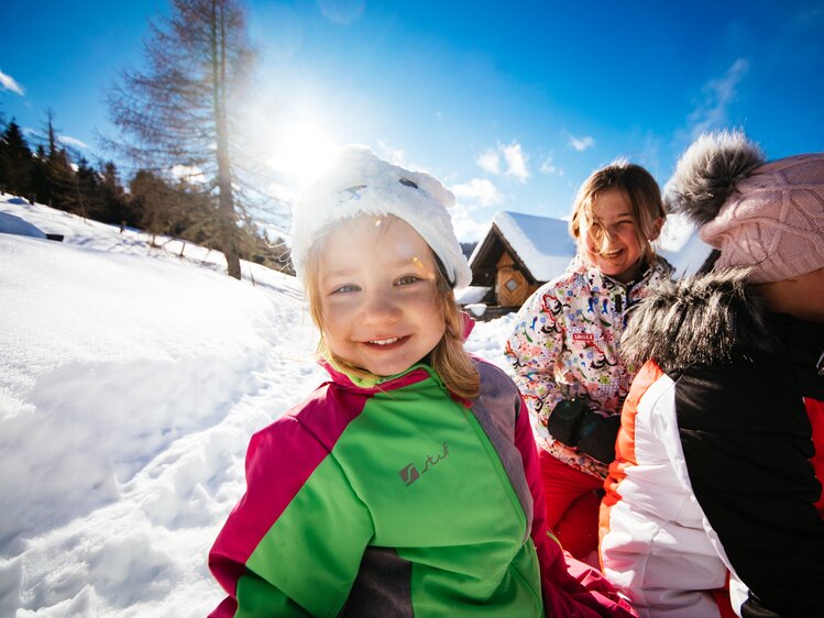 Familienzeit im Winter | © Urlaub am Bauernhof Kärnten / Daniel Gollner Glückliche Kinder, die im Winter beim Schneefall Spaß haben. Sie tragen warme Kleidung, Schal und Mütze, und lächeln in die Kamera. Im Hintergrund sind verschneite Berge und Häuser zu erkennen. | © Urlaub am Bauernhof Kärnten / Daniel Gollner