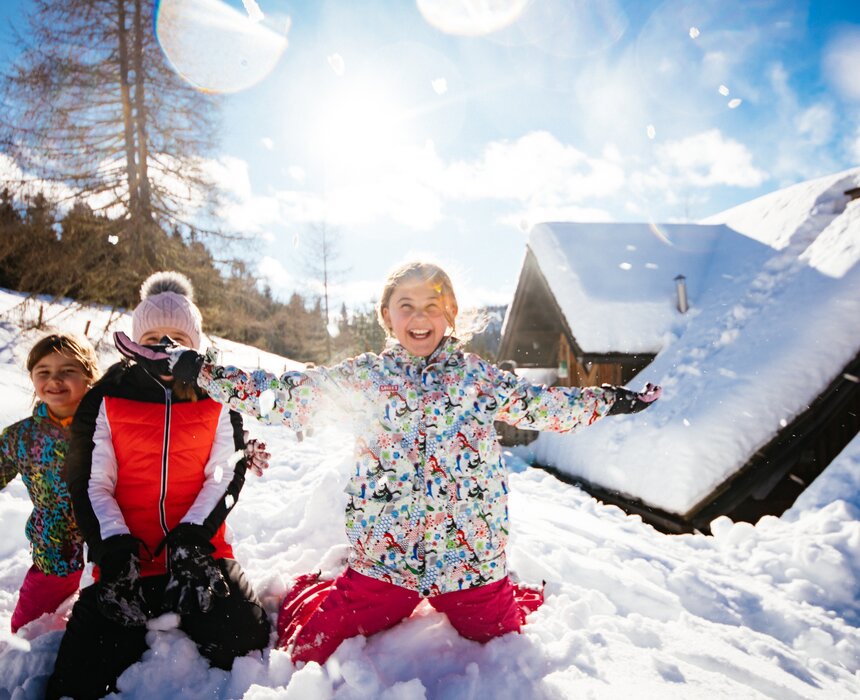 Fröhliche Kinder in bunten Winterjacken und Hosen spielen ausgelassen im tiefen Schnee vor einer verschneiten Berglandschaft, die Arme ausgebreitet vor Freude. | © Urlaub am Bauernhof Kärnten / Daniel Gollner