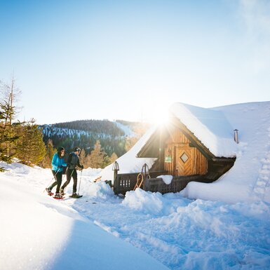 Ein schneebedecktes Berghaus in einer winterlichen Landschaft. Zwei Menschen in Sportkleidung genießen die Winterlandschaft. Die Sonne scheint durch die Bäume und wirft einen warmen Lichtschein auf die Szenerie. | © Daniel Gollner
