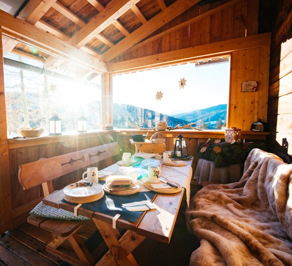 Gemütliche Almhütte mit Holzvertäfelung, großen Fenstern und Blick auf winterliche Berglandschaft. Tisch mit Tellern, Tassen und Kerzen, kuschelige Decke auf Sitzbank. | © Daniel Gollner / Urlaub am Bauernhof Kärnten 