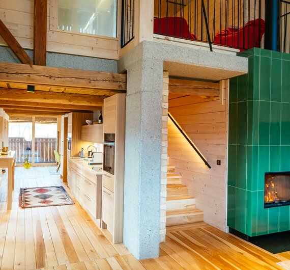 Geräumiges, hölzernes Chalet mit offener Küche, bequemer Sitzecke und farbenfroher Holzofen. Große Fenster bieten Einblick in die gemütliche Atmosphäre. | © Urlaub am Bauernhof Kärnten / Daniel Gollner