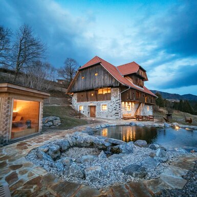 Kaiser Chalet in Kärnten am Abend | © Urlaub am Bauernhof Kärnten / Daniel Gollner Rustikales Blockhaus am Berghang mit beleuchteter Terrasse und Steinpool, umgeben von Bäumen und bewölktem Himmel. | © Urlaub am Bauernhof Kärnten / Daniel Gollner