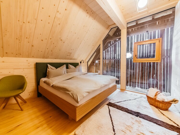 Gemütliches Dachzimmer mit Holzpaneelen, bequemem Doppelbett, Sesseln und Korbmöbeln. Großes Fenster mit Blick nach draußen. Raum erscheint hell und modern eingerichtet. | © Urlaub am Bauernhof Kärnten / Daniel Gollner