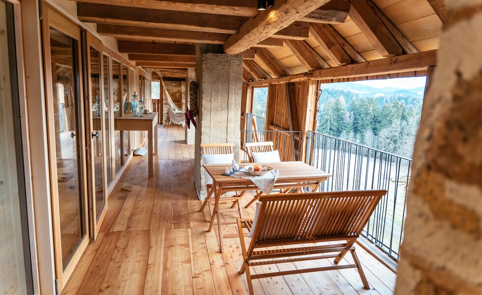 Eine geräumige Holzhütte mit großen Fenstern, die einen Panoramablick auf eine bewaldete Berglandschaft bietet. Innen befinden sich Holztische, Stühle und eine gemütliche Terrasse, die zum Verweilen einladen. | © Urlaub am Bauernhof Kärnten / Daniel Gollner