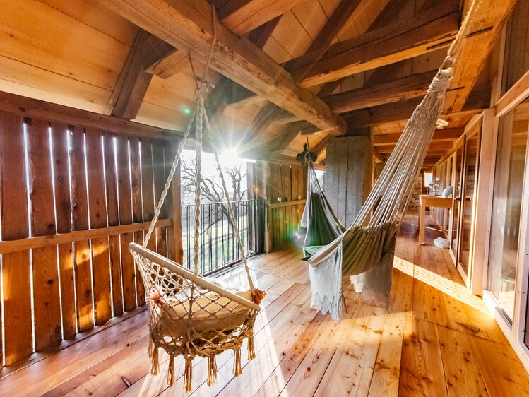 Gemütlicher Holzbalkon mit Hängesessel und Hängematte, umgeben von rustikalen Holzbalken und Fenstern, die einen Ausblick auf eine natürliche Landschaft gewähren. | © Urlaub am Bauernhof Kärnten / Daniel Gollner