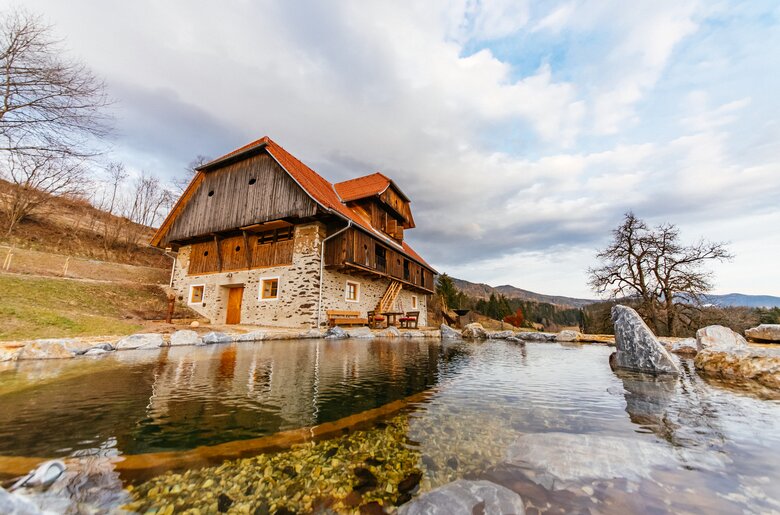 Ein malerisches Gebirgshaus mit Holzfassade und Strohdach direkt am Wasser, umgeben von Bäumen und Felsen. Ein ruhiger See spiegelt die Gebäude und die Wolken am Himmel wider. Die Szene strahlt Natürlichkeit und Ruhe aus. | © Urlaub am Bauernhof Kärnten / Daniel Gollner