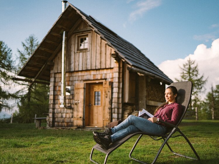 Eine Person sitzt in einem Campingstuhl vor einer alten, holzverkleideten Hütte in einer ländlichen Umgebung und genießt die Natur, während sie ein Buch liest. | © Urlaub am Bauernhof / Daniel Gollner