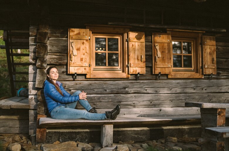 Eine Frau entspannt sich vor der Kleinen Almhütte | © Urlaub am Bauernhof / Daniel Gollner Eine Frau in blauer Jacke sitzt auf einer Holzbank vor einer rustikalen Hütte mit Fensterläden. Die Hütte ist von Holz und befindet sich in einer natürlichen Umgebung. | © Urlaub am Bauernhof / Daniel Gollner