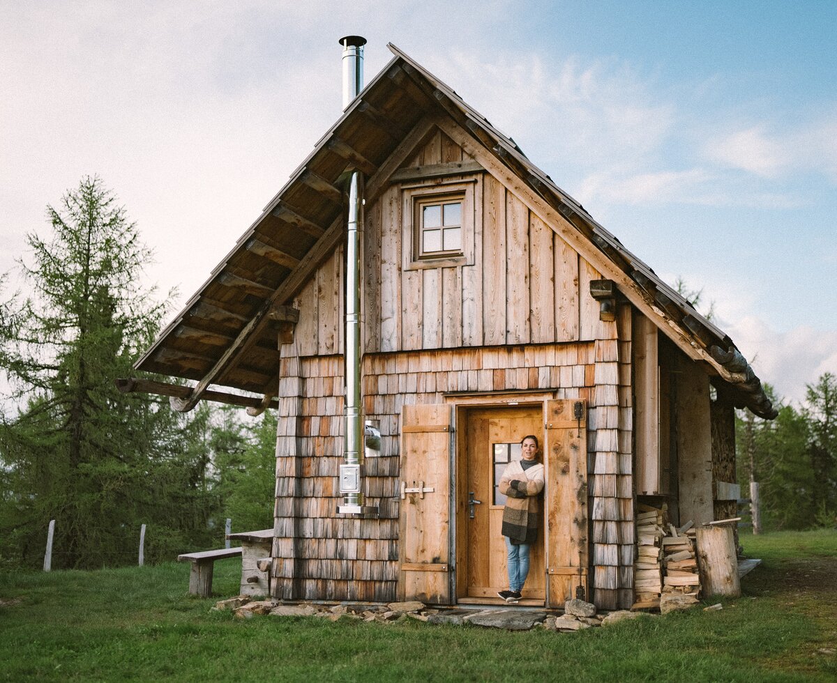 Eine Frau steht in der Tür der Kleinen Almhütte | © Urlaub am Bauernhof / Daniel Gollner Eine rustikale Holzhütte mit Schindeldach umgeben von Bäumen. Eine Person steht in der Tür der Hütte. Die Hütte wirkt ländlich und naturnah in einer waldähnlichen Umgebung. | © Urlaub am Bauernhof / Daniel Gollner