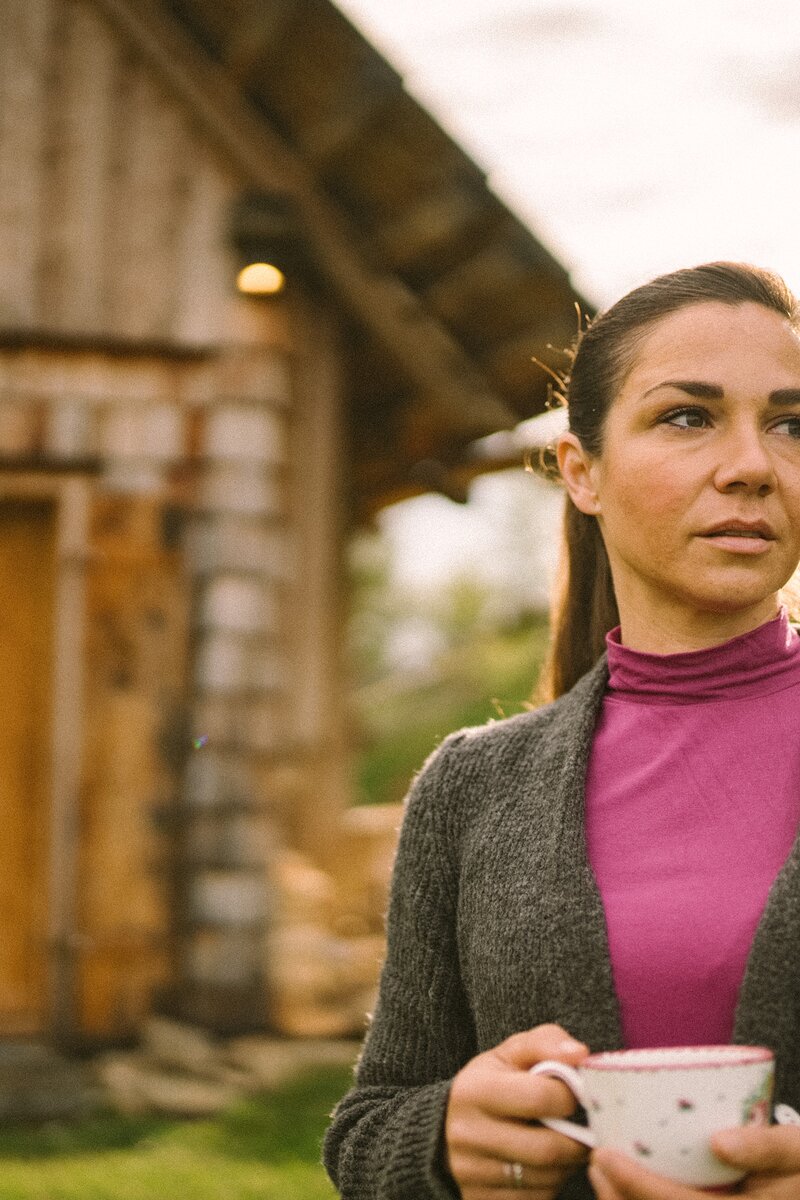 Eine Frau hält eine Teetasse in ihren beiden Hände. Im Hintergrund erkennt man unscharf die Kleine Almhütte. Die Sonne scheint auf den Rücken der Frau.  | © Urlaub am Bauernhof / Daniel Gollner