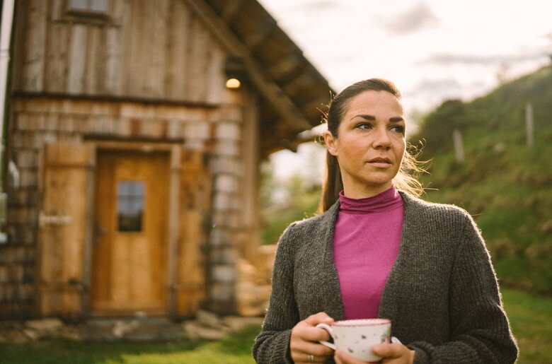 Eine Frau in einem grauen Pullover und einer rosa Jacke steht vor einer alten Holzhütte auf einer Wiese und hält eine Tasse in der Hand. Windspiele treiben in der Luft, was eine ruhige Atmosphäre vermittelt. | © Urlaub am Bauernhof / Daniel Gollner
