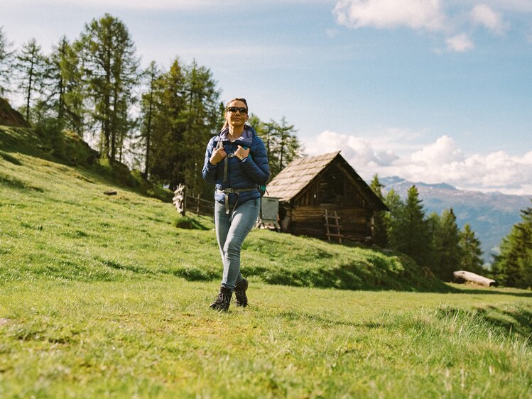 Eine Wanderin in Jeans und Jacke steht auf einer grünen Wiese vor einer Holzhütte, umgeben von Bäumen und Berglandschaft in der Ferne. | © Urlaub am Bauernhof Kärnten / Daniel Gollner