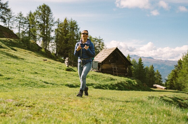 Eine Wanderin in Jeans und Jacke steht auf einer grünen Wiese vor einer Holzhütte, umgeben von Bäumen und Berglandschaft in der Ferne. | © Urlaub am Bauernhof Kärnten / Daniel Gollner