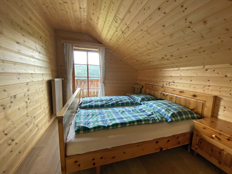 Dies ist ein Schlafzimmer in einer gemütlichen Berghütte. Der Raum ist aus hellem Holz gebaut, mit einem bequemen Doppelbett, einem Fenster und rustikalen Möbeln. Die karierte Bettwäsche verleiht dem Raum einen ländlichen, bergigen Charakter. | © Jasmin Kreulitsch