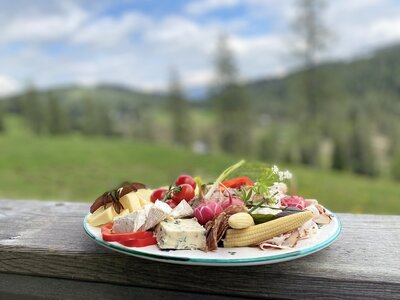 Brettljause Mechtenhütte | © Urlaub am Bauernhof Kärnten / Jasmin Kreulitsch Eine Auswahl an Wurst, Käse, Tomaten, Gewürzen und anderen Lebensmitteln auf einem holzigen Untergrund, umgeben von einer grünen, bergigen Landschaft. | © Urlaub am Bauernhof Kärnten / Jasmin Kreulitsch