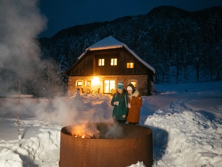 Feuerschale vor der Obiralmhütte | © Urlaub am Bauernhof / Daniel Gollner Eine verschneite Hütte bei Nacht, mit warm erleuchteten Fenstern und einem lodernden Feuer im Freien, an dem zwei Menschen stehen und sich wärmen. | © Urlaub am Bauernhof / Daniel Gollner