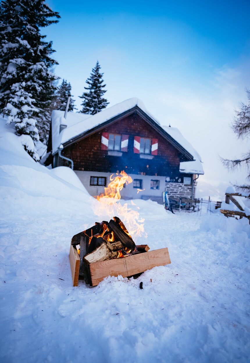 Ein schneebedecktes Holzhaus mit brennendem Kamin vor einer Berglandschaft. Davor liegt gehacktes Brennholz in einem hölzernen Gestell, umgeben von tiefer Schneedecke. | © Urlaub am Bauernhof Kärnten/ Daniel Gollner