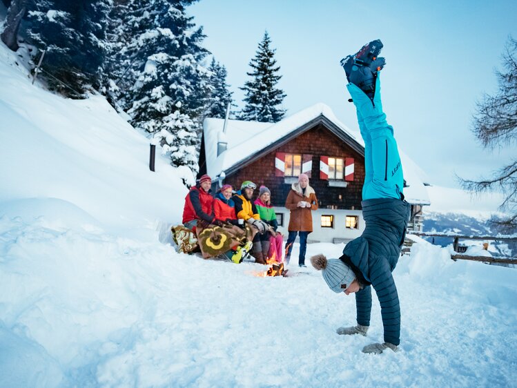 Frau macht einen Handstand im Schnee | © Urlaub am Bauernhof Kärnten/ Daniel Gollner Gruppe von Wintersportlern vor einer gemütlichen Hütte im verschneiten Wald, eine Person macht einen Handstand im Schnee. | © Urlaub am Bauernhof Kärnten/ Daniel Gollner