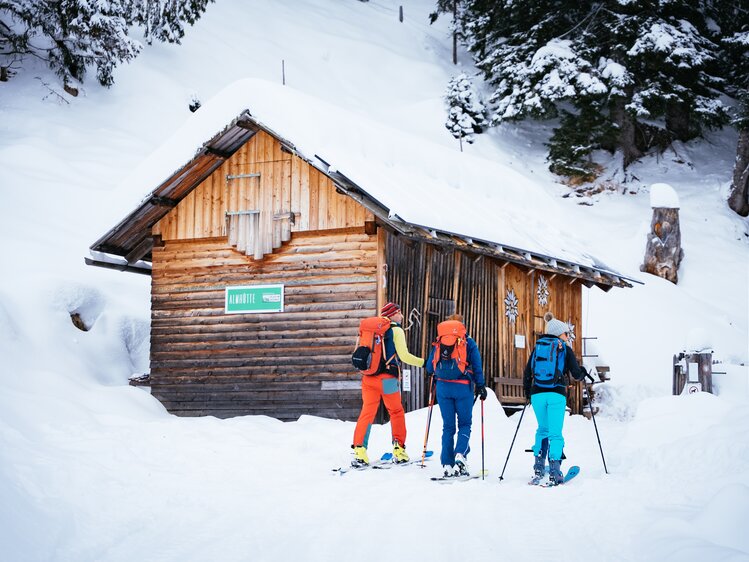 Freunde machen eine Skitour | © Urlaub am Bauernhof Kärnten/ Daniel Gollner Eine Holzhütte inmitten verschneiter Wälder, vor der drei Skifahrer in bunter Winterkleidung stehen. Die Szene vermittelt Entspannung und den Genuss der winterlichen Natur. | © Urlaub am Bauernhof Kärnten/ Daniel Gollner