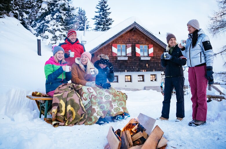 Freunde sitzen bei der Feuerstelle | © Urlaub am Bauernhof Kärnten/ Daniel Gollner Gruppe von Freunden, die bei einem verschneiten Berghüttchen eine gemütliche Winterrast machen. Sie sitzen um ein Lagerfeuer herum und genießen heiße Getränke. Die Hütte im traditionellen Holzbau-Stil liegt inmitten einer idyllischen, winterlichen Landschaft. | © Urlaub am Bauernhof Kärnten/ Daniel Gollner