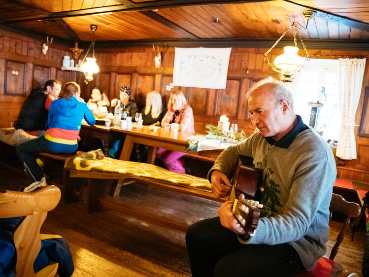 Eine gemütliche Holzhütte mit einer Gruppe von Menschen, die gemeinsam essen und trinken. In der Ecke sitzt ein älterer Mann, der Gitarre spielt. An den Wänden hängen Jagdtrophäen und andere dekorative Elemente, die eine traditionelle Alpine-Atmosphäre schaffen. | © Urlaub am Bauernhof Kärnten/ Daniel Gollner