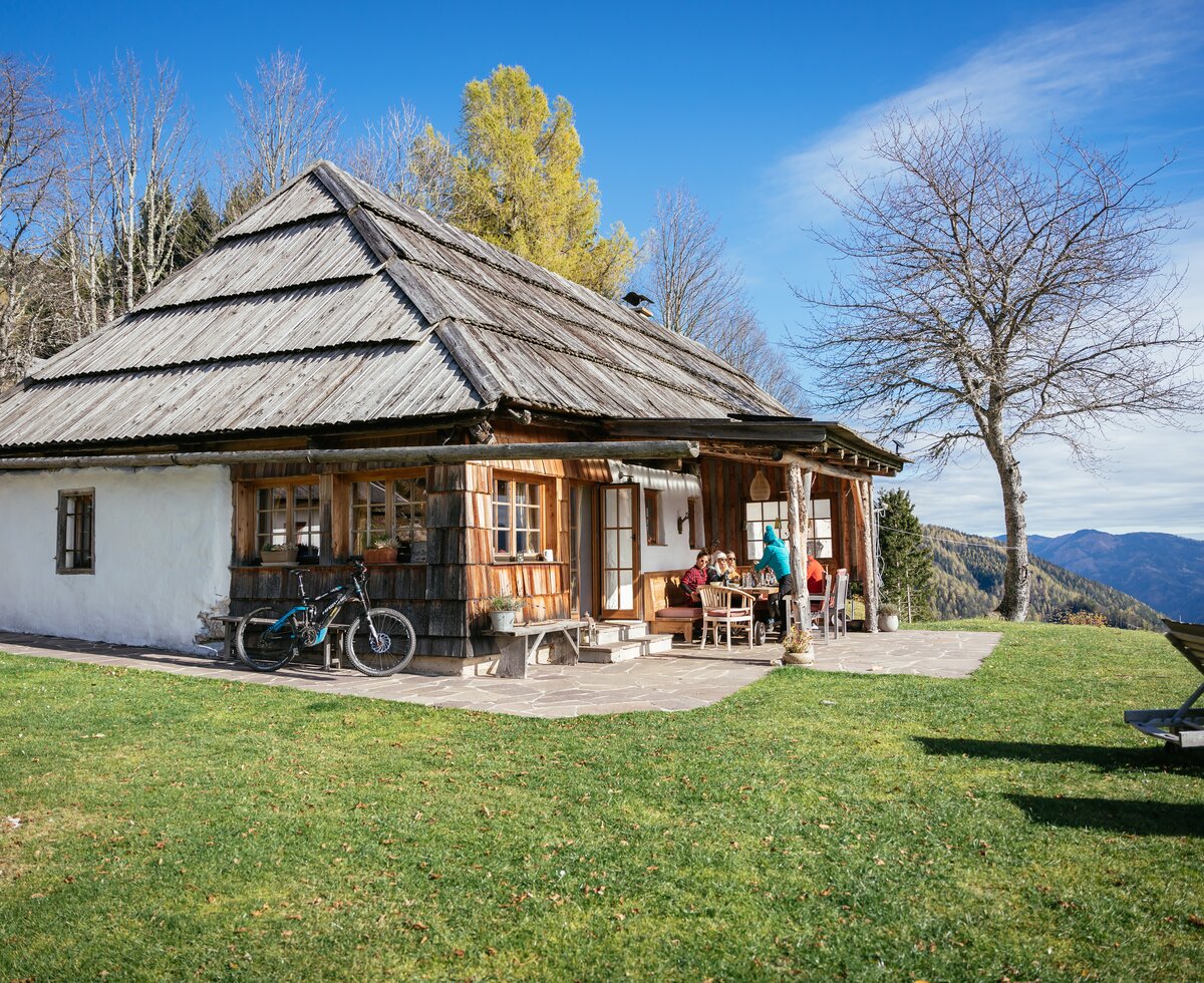 Obiralmhütte Meierhof | © Urlaub am Bauernhof Kärnten/ Daniel Gollner Eine traditionelle Hütte mit Holzdach und Veranda auf einer Wiese mit Wald und Bergen im Hintergrund. Ein Fahrrad und eine Liege sind vor dem Haus zu sehen. | © Urlaub am Bauernhof Kärnten/ Daniel Gollner