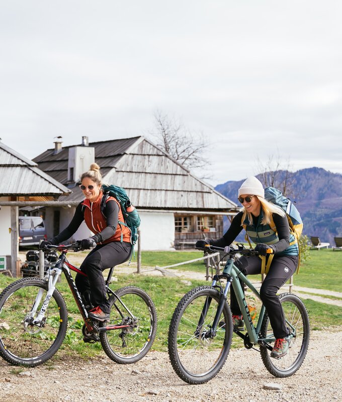 Zwei Frauen mit Fahrrädern und Rucksäcken vor einem ländlichen Gebäudekomplex mit Holzdächern. Im Hintergrund sind bergige Landschaften zu sehen. | © Urlaub am Bauernhof Kärnten/ Daniel Gollner