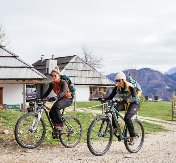 Zwei Frauen mit Fahrrädern und Rucksäcken vor einem ländlichen Gebäudekomplex mit Holzdächern. Im Hintergrund sind bergige Landschaften zu sehen. | © Urlaub am Bauernhof Kärnten/ Daniel Gollner