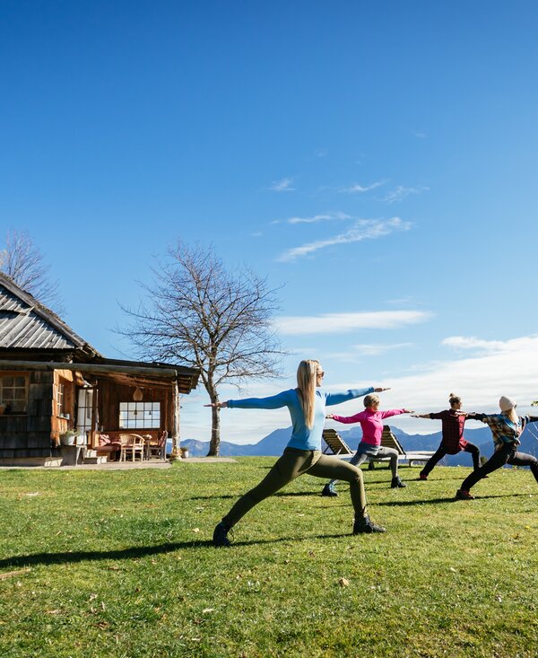Eine Gruppe von Personen macht Yoga-Übungen auf einer Wiese vor einer traditionellen Holzhütte mit Bergkette im Hintergrund. | © Urlaub am Bauernhof Kärnten/ Daniel Gollner